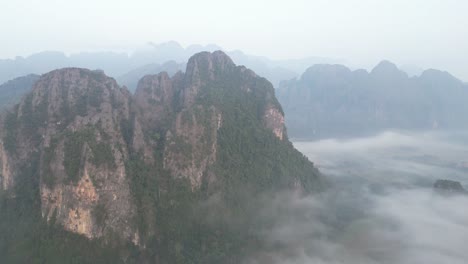Drone-shot-flying-over-cliffs-and-misty-valley-in-Vang-Vieng,-the-adventure-capital-of-Laos