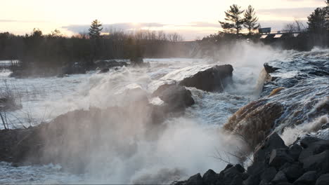 Misty-waterfalls-cascade-over-rocks-at-sunset,-the-industrial-silhouette-of-a-hydropower-plant-in-the-background
