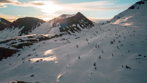 Snow-covered-mountains-bathed-in-warm-sunset-light,-aerial-shot-of-serene-alpine-landscape