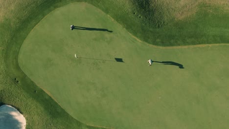 Top-down-bird's-eye-view-of-two-male-golfers-and-a-flag-stick-with-long-shadows-putting-a-golf-ball-into-the-hole-on-a-sunny-afternoon-on-a-Florida-golf-course