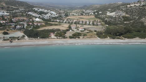 Toma-Aérea-De-La-Playa-De-Pissouri-En-Chipre-Con-Serenas-Aguas-Azules-Y-Fondo-De-Ciudad-Costera,-Luz-Del-Día