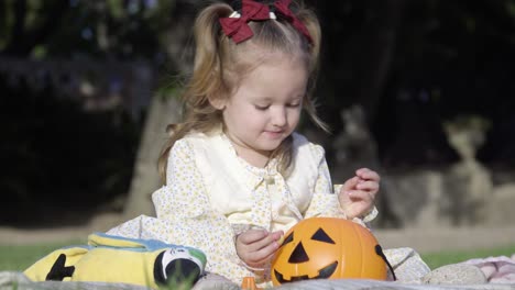 Close-up-of-adorable-little-girl-sitting-on-the-blanket-and-playing-with-the-halloween-pumpkin-in-the-city-park
