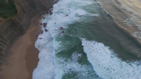 Aerial-view-tilting-over-waves-at-the-Magoito-Beach,-cloudy-sunset-in-Portugal
