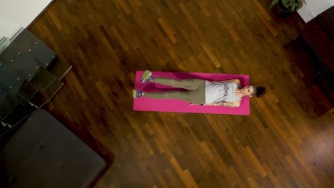 Woman-doing-flutter-kicks-on-a-pink-mat,-hardwood-floor,-top-down-view,-with-a-tattoo