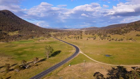 Drone-view-of-car-driving-on-country-road-with-grassy-rural-farmland-on-sunny-day,-Crackenback,-New-South-Wales,-Australia