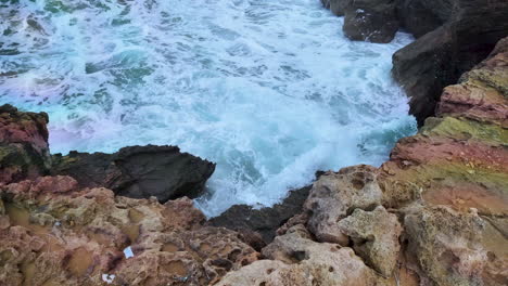 Rocky-coastal-scene-where-turbulent-white-waves-crash-against-jagged,-reddish-brown-rocks