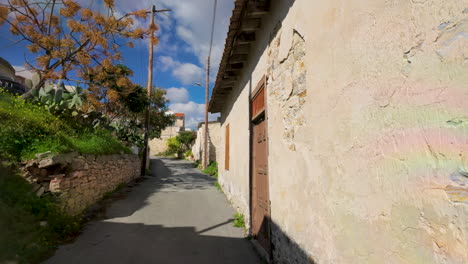 Un-Callejón-Iluminado-Por-El-Sol-En-Lefkara,-Flanqueado-Por-Tradicionales-Paredes-Encaladas,-Una-Antigua-Puerta-De-Madera-Y-Un-árbol-En-Flor,-Que-Evoca-El-Encanto-Rústico-Del-Pueblo