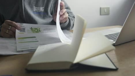 Closeup-shot,-female-elder-hands-go-through-paper-payment-bills,-desktop-office-with-laptop