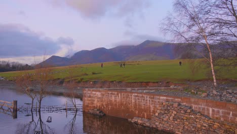 Hikers-walking-near-a-calm-lake-in-Lake-District-at-sunset,-mountain-backdrop