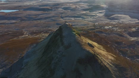 Flying-over-isolated-peak-in-winter-early-morning-with-reveal-of-rugged-moorland,-coastline-and-distant-mountains-beyond