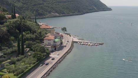 Vista-Aérea-Del-Paseo-Del-Lago-En-El-Lago-De-Garda-Con-Coches-En-La-Carretera-Y-Windsurfistas-En-El-Lago-En-Un-Día-Soleado