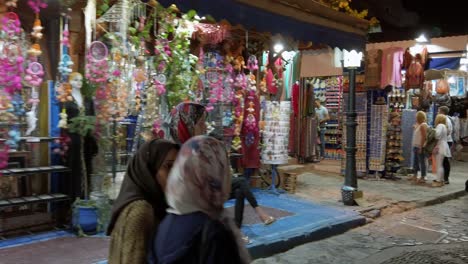 Traditional-Arabic-women-walk-in-front-of-the-vibrant-and-illuminated-souvenir-shops-in-the-centre-of-Chefchaouen-at-night-in-Morocco
