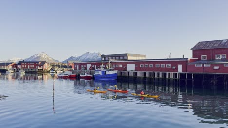 Kayakers-paddle-by-colorful-waterfront-buildings-in-Lofoten-with-mountains-backdrop,-calm-waters