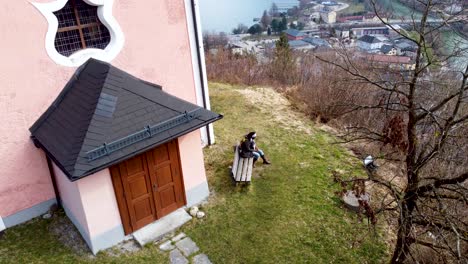 Black-Woman-is-sitting-near-picturesque-chapel-on-Kalvarienberg-overlooking-Traunsee-in-Ebensee,-Upper-Austria,-framed-by-Alps