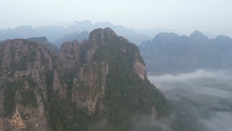 drone-shot-of-dramatic-cliffs-above-foggy-valley-in-Vang-Vieng,-the-adventure-capital-of-Laos