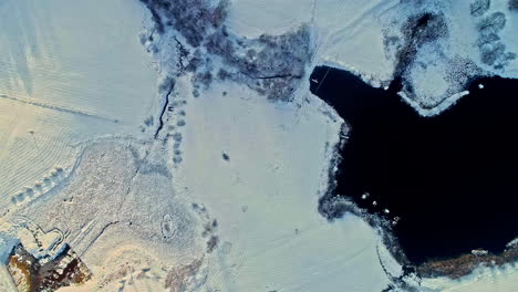 A-snow-covered-landscape-with-contrasting-dark-water-body,-aerial-view