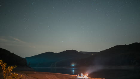 Cielo-Nocturno-Sobre-Un-Lago-Sereno-Con-Estrellas,-Pequeña-Fogata-En-La-Orilla,-Montañas-Al-Fondo,-Atmósfera-Tranquila,-Timelapse