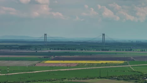 Toma-Aérea-Distante-De-Un-Largo-Puente-Suspendido-Enmarcado-Por-Verdes-Campos-Agrícolas-Y-Un-Cielo-Azul-Nublado