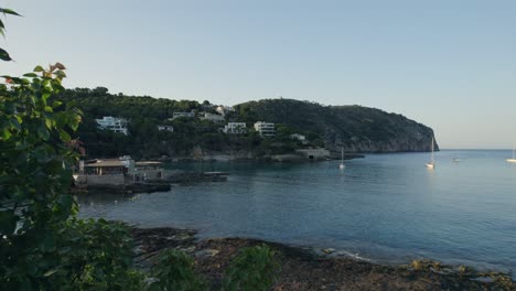 Drone-lifting-from-the-pool-side-to-display-the-beach,-boats-and-hill-in-the-horizon,-Mallorca-Spain