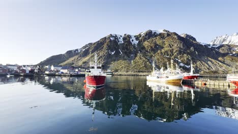 Red-fishing-boats-docked-in-calm-waters-with-snowy-Lofoten-mountains-reflecting-in-Norway,-clear-sky