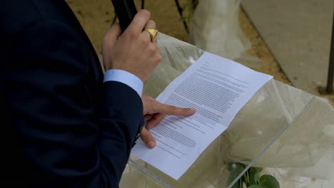 a-man-reads-a-text-on-a-transparent-plexiglass-lectern-at-a-wedding,-it's-a-speech,-focus-on-the-sheet-and-the-finger-running-over-the-lines,-the-hand-holding-the-microphone