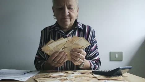 Kaukasische-Frau-Mittleren-Alters-Mit-Weißem-Haar-Zählt-50-Euro-Scheine-Papiergeld,-Desktop-Büro-Mit-Einem-Taschenrechner-Und-Steuern