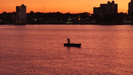 Panoramic-shot-of-a-fisherman-in-a-canoe,-sailing-the-Paraná-River-with-the-city-of-Posadas-at-sunset-in-the-background,-Argentina
