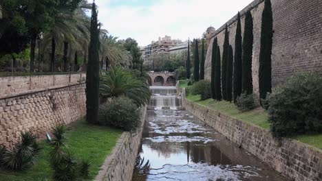 Baluard-de-Sant-Pere-walls,-Riera-river,-bridge-and-cypress-trees-in-Palma-de-Mallorca,-Spain-next-to-the-urban-park