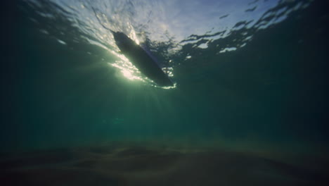 Silhouette-Eines-Longboard-Surfers-Im-Gegenlicht-Der-Morgenstrahlen-Unter-Wasser