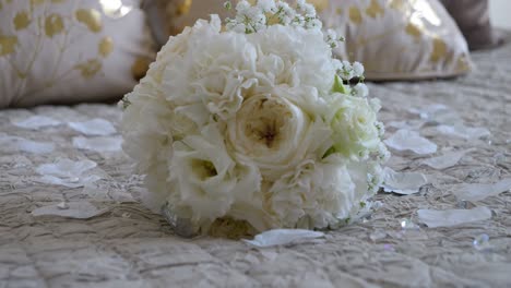 Drehaufnahme-Und-Fokus-Auf-Den-Weißen-Blumenstrauß-Der-Braut-Auf-Einem-Luxuriösen-Bett-Mit-Weißen-Blütenblättern-Rundherum,-Weiße-Und-Goldene-Bettwäsche