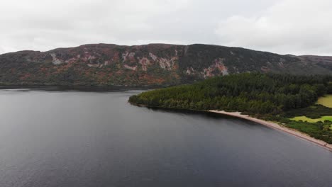 Aerial-View-Of-Beach-Shoreline-At-Loch-Ness-With-Mountain-Landscape-In-Background