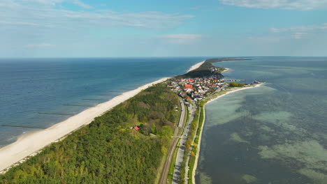 Vista-Aérea-De-Una-Estrecha-Franja-De-Tierra-Con-Una-Carretera-Que-Divide-La-Densa-Vegetación-Y-Un-Pueblo-Costero-De-La-Extensa-Playa-De-Arena-Y-El-Mar-Abierto-Por-Un-Lado-Y-La-Tranquila-Bahía-Por-El-Otro---Kuźnica,-Hel