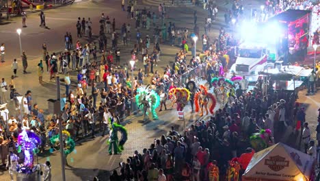 Feathery-costume-performers-dance-and-sway-at-night-illuminated-by-lights,-aerial-orbit