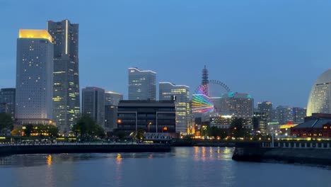 Abendlicher-Blick-Auf-Die-Lebendige-Skyline-Der-Stadt-Mit-Beleuchteten-Gebäuden-Und-Einem-Bunten-Riesenrad,-Das-Sich-Im-Wasser-Spiegelt