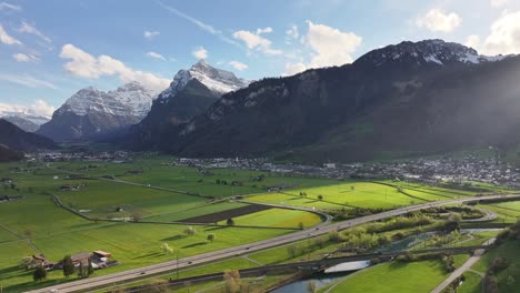 Aerial-of-Lush-green-valley-near-Walensee,-Weesen,-Switzerland,-with-sprawling-fields-and-majestic-snow-capped-mountains-under-clear-sky