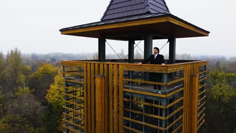 Hombre-De-Abrigo-Negro-Parado-En-La-Torre-De-Vigilancia-Sobre-El-Bosque,-Establecimiento-Aéreo