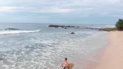 Drone-Flying-Over-Shirtless-Man-Running-On-Sandy-Beach-With-Surfboard-in-Sri-Lanka