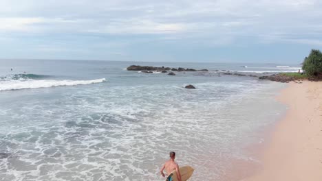 Drone-Volando-Sobre-Un-Hombre-Sin-Camisa-Corriendo-En-Una-Playa-De-Arena-Con-Tabla-De-Surf-En-Sri-Lanka