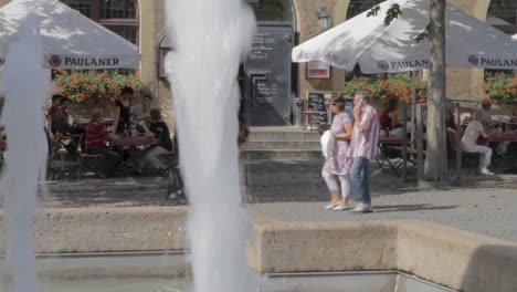 Cafe-scene-with-people-dining-under-umbrellas,-viewed-past-a-fountain-on-a-sunny-day