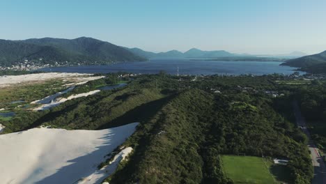 A-drone-advances-over-the-sand-dunes-of-Joaquina,-unveiling-the-vast-Lagoa-da-Conceição-in-the-background