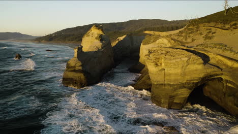 Golden-light-glowing-on-sandstone-cliffs-as-waves-crash-on-hidden-cove,-aerial