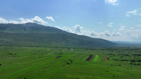 Sweeping-aerial-shot-of-an-expansive-mountain-plateau,-lightly-covered-in-springtime-grass-and-sparse-woodland,-with-the-backdrop-of-high-mountain-peaks-and-a-sky-speckled-with-clouds