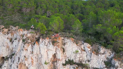 Hikers-rest-on-a-rocky-cliff-among-trees-with-a-view-of-the-green-landscape-during-sunset-in-ibiza