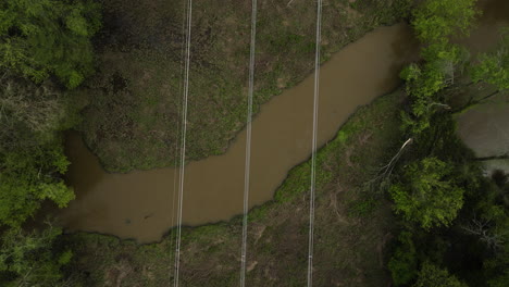 Power-lines-crossing-a-natural-area-with-dense-greenery-and-water-in-tennessee,-aerial-view