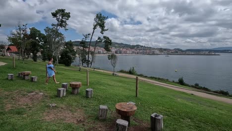 La-Mujer-Sale-A-Disfrutar-De-La-Terraza-De-Césped-Natural-Con-Asientos-Y-Mesas-Rústicas-De-Madera-Frente-Al-Mar-Y-La-Ciudad-Se-Ve-Al-Fondo,-Día-Nublado,-Foto-Descriptiva