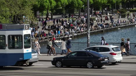 Langsame,-Nach-Links-Fahrende-Drohnenaufnahme-Mit-Der-Zürcher-Tram,-Die-Nach-Links-Aus-Dem-Bild-Fährt-Und-Menschenmengen-Zeigt,-Die-Am-Seeufer-Der-Stadt-Sitzen