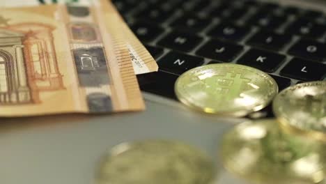 Rotating-close-up-above-Bitcoin-Coins,-50-Euro-paper-bills,-laptop-in-home-desk-office,-digital-versus-paper-currency