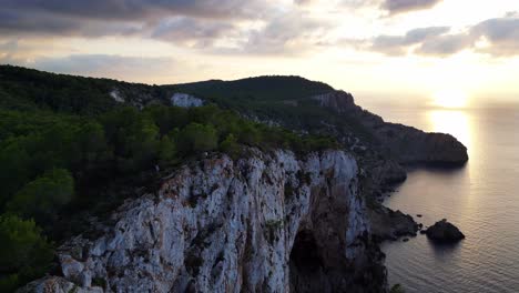 Hikers-rest-on-a-rocky-cliff-among-trees-with-a-view-of-the-green-landscape-during-sunset-in-ibiza