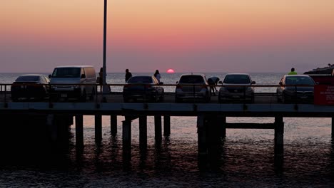 Silhouette-Von-Autos,-Die-Auf-Dem-Pier-Geparkt-Sind,-Während-Menschen-Bilder-Vom-Sonnenuntergang-Unter-Dem-Horizont-Am-Meer-Machen