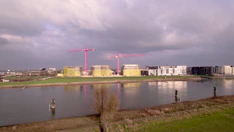 Aerial-river-IJssel-passing-by-Dutch-new-build-neighbourhood-Noorderhaven-with-red-cranes-rising-above-construction-site